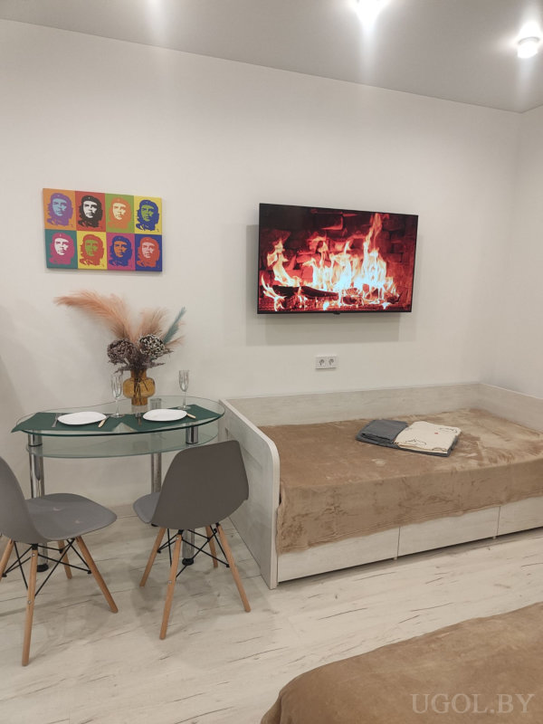 Настенный телевизор с изображением камина, диван-деньбед с покрывалом, стеклянный стол на двоих с сервировкой и вазой с сухоцветами, над столом яркая поп-арт картина.