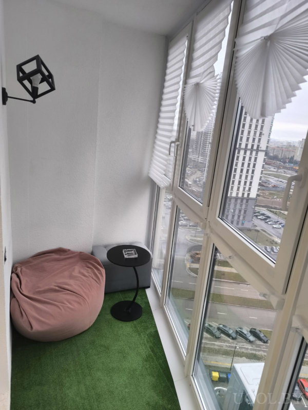 Застеклённая лоджия с зелёным ковром, розовым креслом-мешком и столиком с пепельницей, рядом серый пуф, на стене светильник, за окнами вид на многоэтажные дома и улицы города.