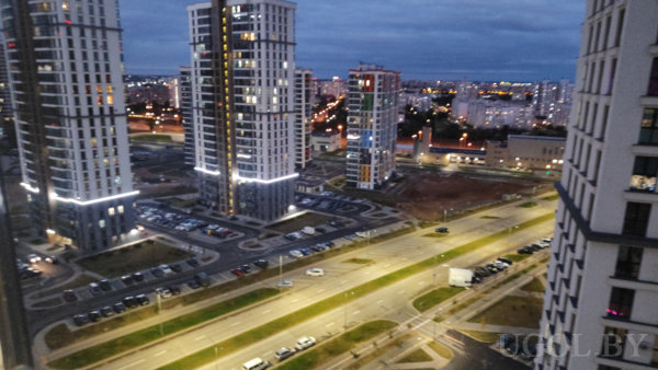 Современные многоэтажные дома с подсветкой и большими окнами, освещённые улицы с машинами и парковками, зелёные зоны вдоль дороги, вдалеке огни густонаселённого района.