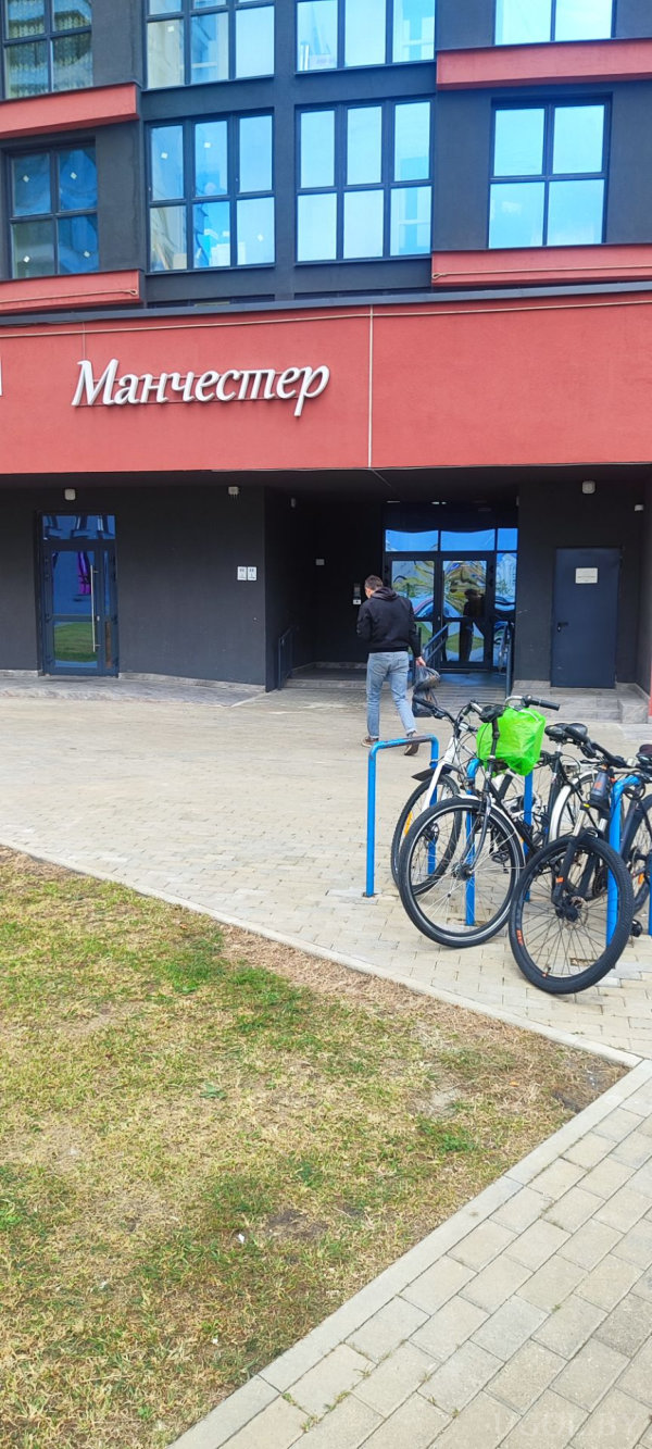 Современный фасад с тёмной отделкой и большими окнами, над входом вывеска «Манчестер», рядом велопарковка с велосипедами и человек у домофона.