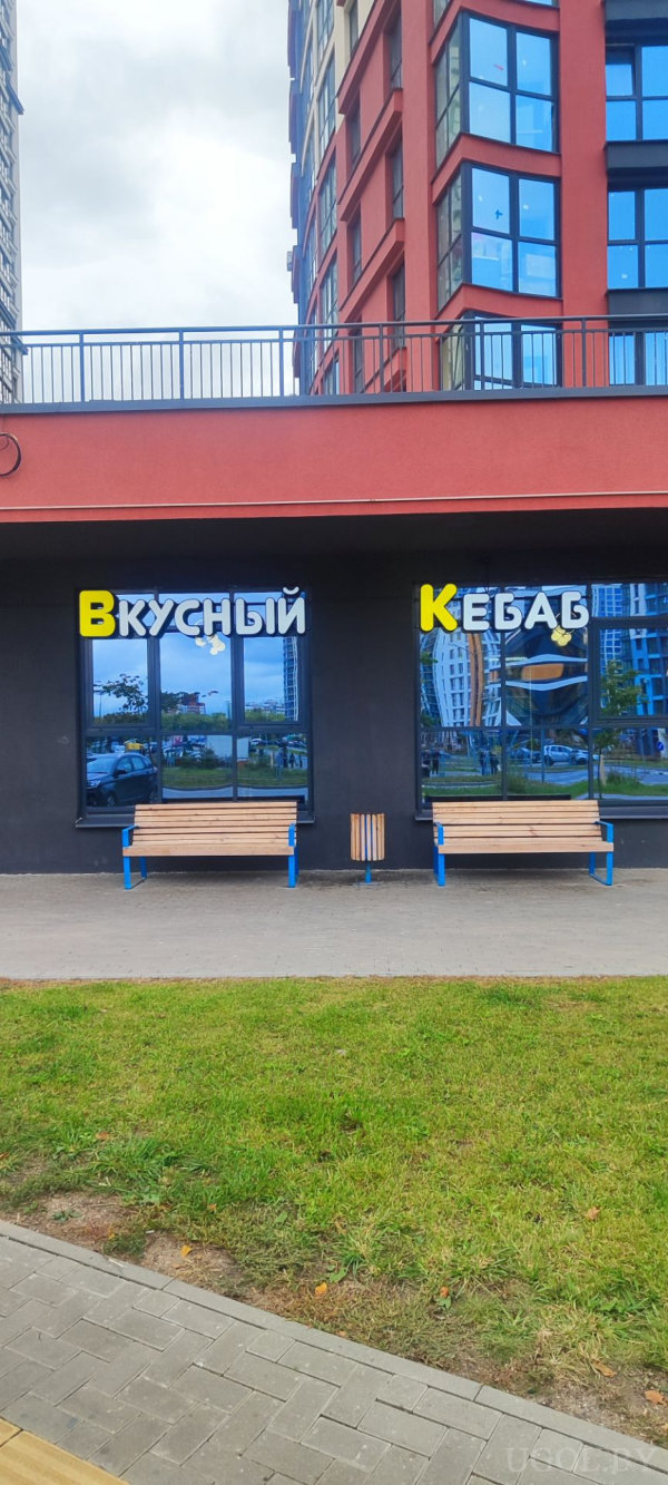 Красный фасад здания с большими окнами, вывеска «Вкусный Кебаб», перед входом две деревянные лавочки с синими металлическими опорами и урна, аккуратная городская среда.