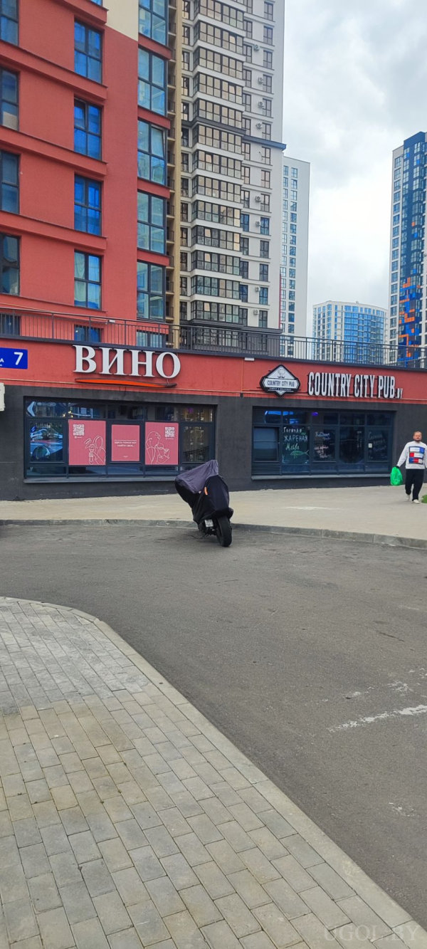 Современные жилые дома с красными и нейтральными фасадами, на первом этаже магазин с вывеской «Вино» и заведение «Country City Pub», рядом припаркованный мотоцикл и прохожий с зелёной сумкой.