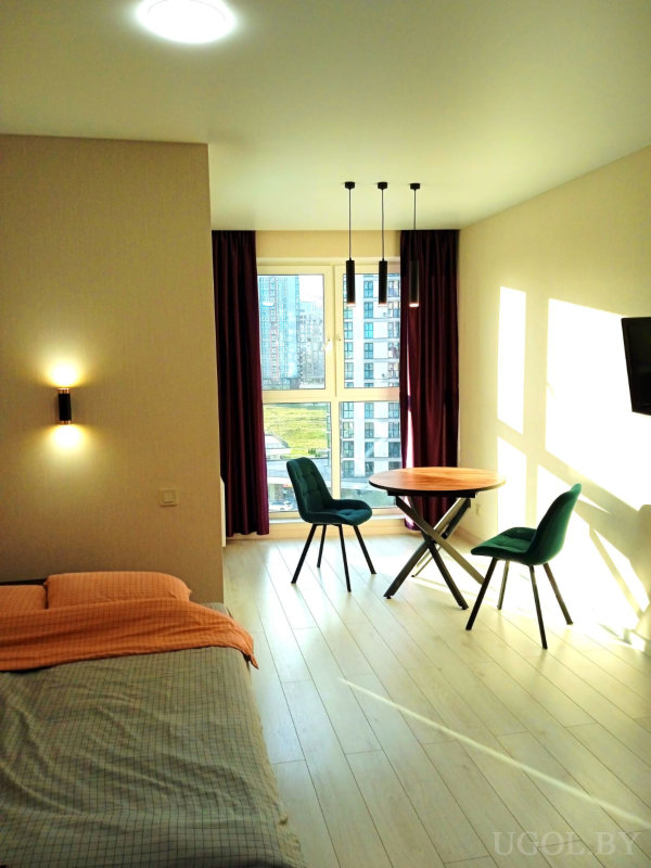 Кровать с серо-оранжевым покрывалом, круглый стол с зелёными креслами, три подвесных светильника и окно с бордовыми шторами.