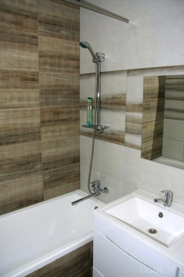 Белая ванна, душевая стойка, стеклянная полка с флаконом, тумба с раковиной и плитка с древесной текстурой.