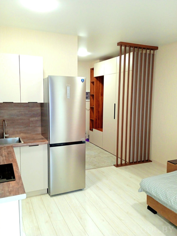 Кухня с белыми фасадами, деревянная перегородка, встроенный шкаф, кровать с серым покрывалом и машинка в нише.