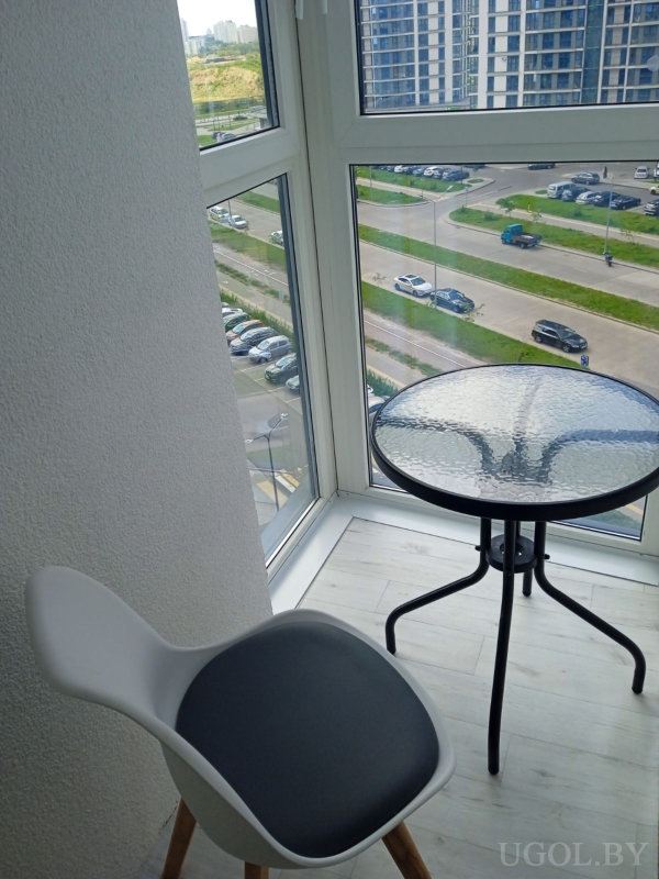 Стеклянный стол с чёрными ножками, белый стул с серой подушкой, светлый пол и панорамные окна с видом на дома и парковку.
