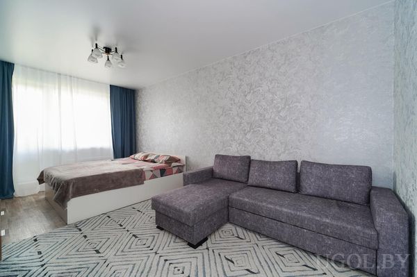 Минималистичная комната с серым диваном, кроватью с коричневым покрывалом, светлым ковром и синими шторами.