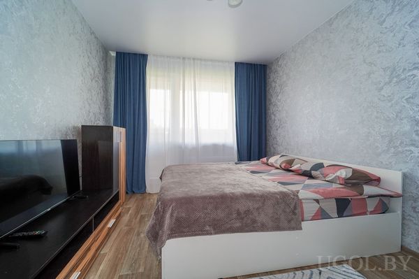 Светлая спальня с кроватью у стены, коричневым покрывалом, телевизором на тумбе и ламинатом на полу.