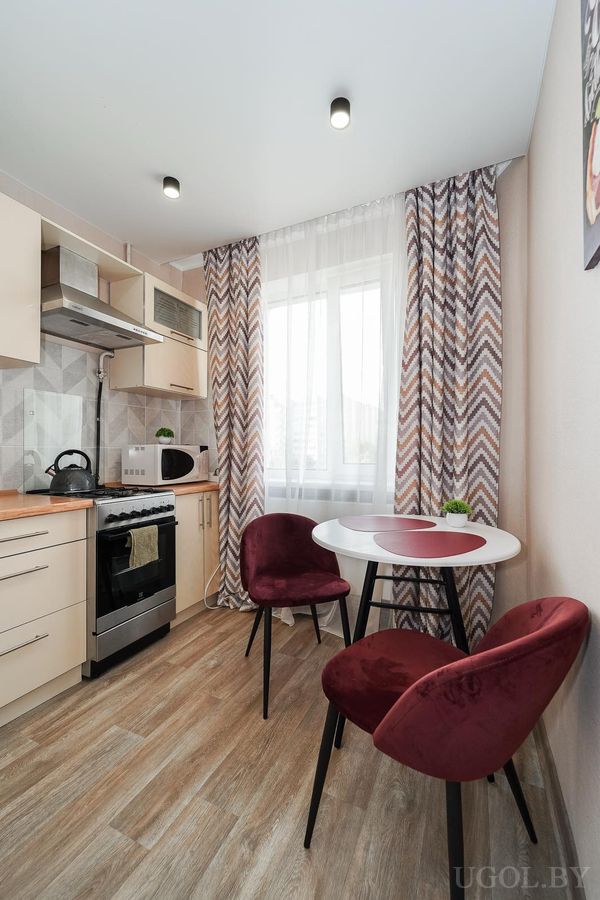 Небольшая кухня с белым столом, бордовыми стульями, плитой и шторами с геометричным узором.