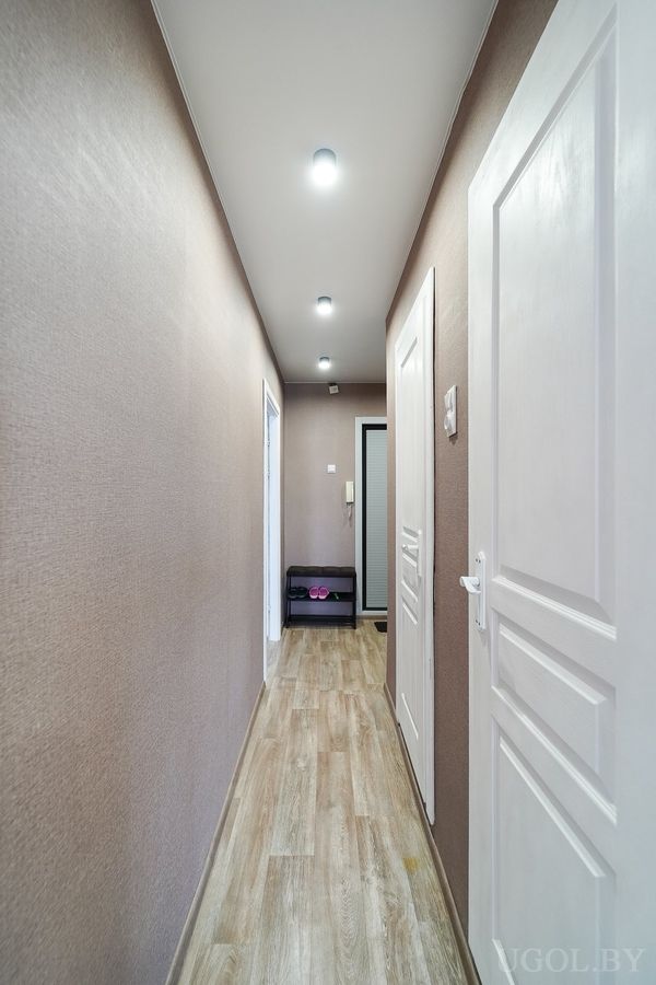 Узкий коридор с белыми дверями, светлым полом, обувной полкой и домофоном на стене.