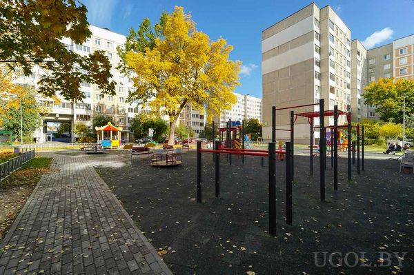 Площадка с горкой, турниками, резиновым покрытием и жёлтыми листьями на фоне многоэтажек.
