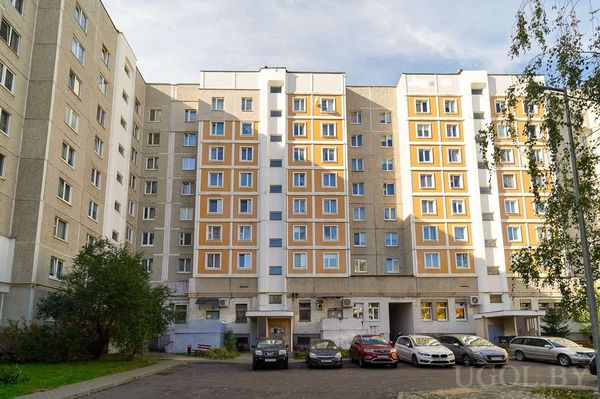 Жилой дом с серым фасадом, оранжевыми акцентами, машинами на парковке и облачным небом.