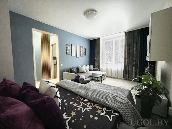 Кровать со звёздным покрывалом, диван, кофейный столик, лампа и синяя стена с картинами, окно с тюлем и портьерами.