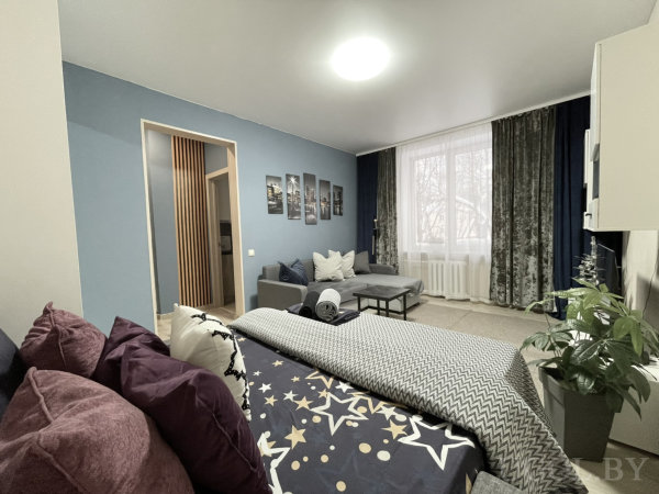 Серый диван, кофейный столик, серия городских фото на синей стене, крупное окно и зелёные растения создают уют.