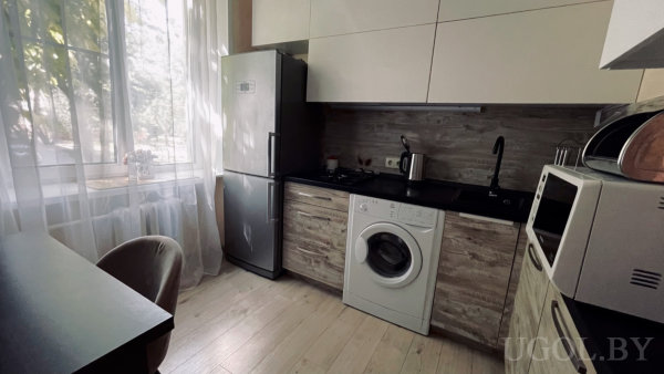 Окно с тюлем, холодильник, мойка, чайник, стиральная машина, микроволновка и часть обеденного стола.