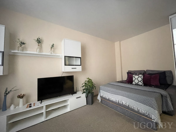 Серая кровать с пледом, белая ТВ-тумба, полка с стеклянными вазами, навесной шкаф и высокое растение — компактный уют.