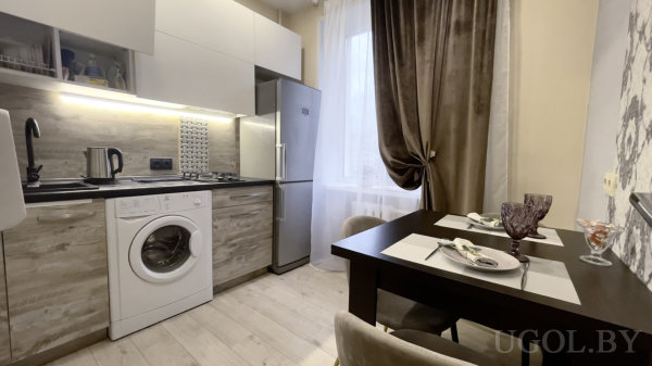 Стиральная машина под столешницей, газовая плита, холодильник, белые и деревянные фасады, сервировка на двоих у окна.