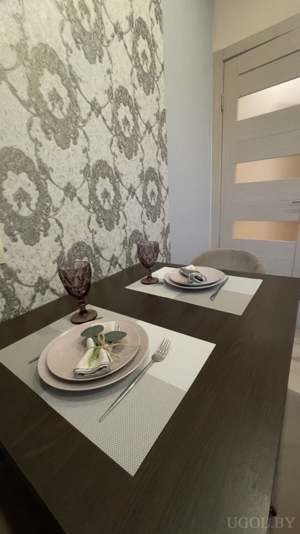 Тёмный стол, светлые тарелки с свёрнутыми салфетками и зеленью, фиолетовые бокалы и орнаментальная серая стена.