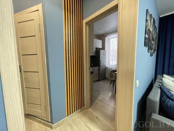 Светлый коридор, декоративные вертикальные панели, кухня с белыми шкафами и окно, вид на гостиную с синей стеной.