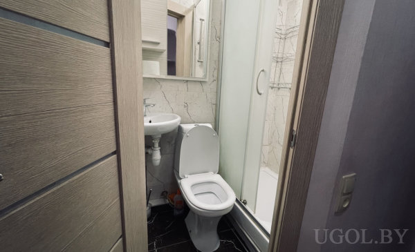 Белый унитаз, маленькая подвесная раковина, стеклянная душевая с раздвижной дверью, светлые стены под мрамор и тёмный глянцевый пол.