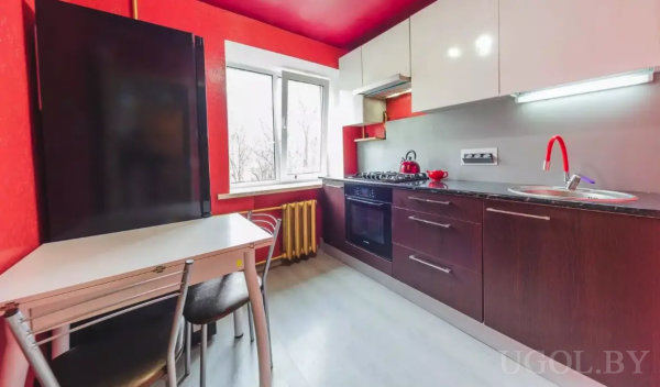 Компактная кухня с белыми и темными шкафами, красным чайником, газовой плитой и обеденной зоной у окна.