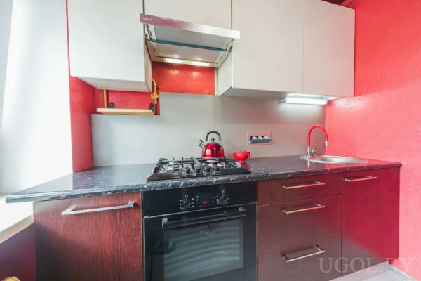 Черная столешница, красный смеситель, белые верхние шкафы и вытяжка над плитой на фоне красных стен.