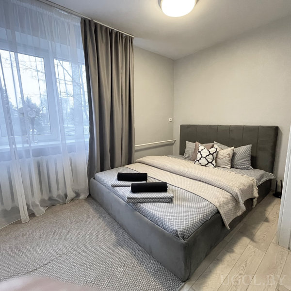 Кровать с серым изголовьем, серо-белым бельём и тремя свернутыми полотенцами, окно с тюлем и шторами.