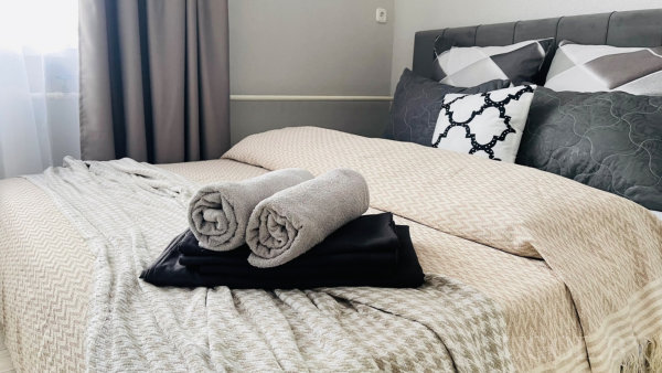 Серое изголовье, бежевое покрывало, декоративные подушки и полотенца, окно с серыми шторами.