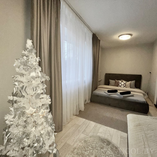 Кровать с подушками и полотенцами, белая искусственная ёлка, круглый ковёр и бежевые шторы.