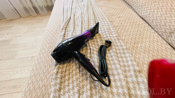 Чёрно-фиолетовый фен с насадкой на светлом пледе, деревянный пол и занавеска на фоне.