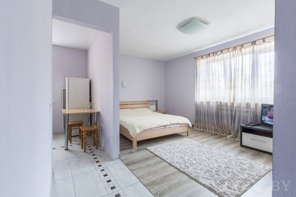 Кровать у стены лавандового оттенка, напротив — телевизор, рядом стол со стульями на плитке с бордюром. Пространство оформлено в мягких тонах и наполнено светом.