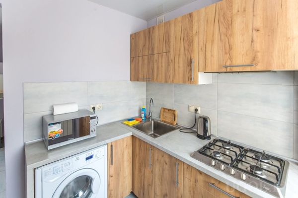 Компактная кухня с деревянными фасадами, плитой, микроволновкой, чайником и стиральной машиной. Светло-серый фартук и столешница подчеркивают аккуратность.