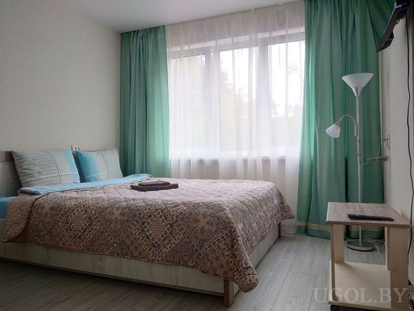 Светлая спальня с большой кроватью в коричневом покрывале, голубыми подушками и телевизором на стене.