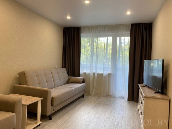 Современная гостиная с бежевым диваном и креслом, телевизором на тумбе и большим окном с видом на деревья.