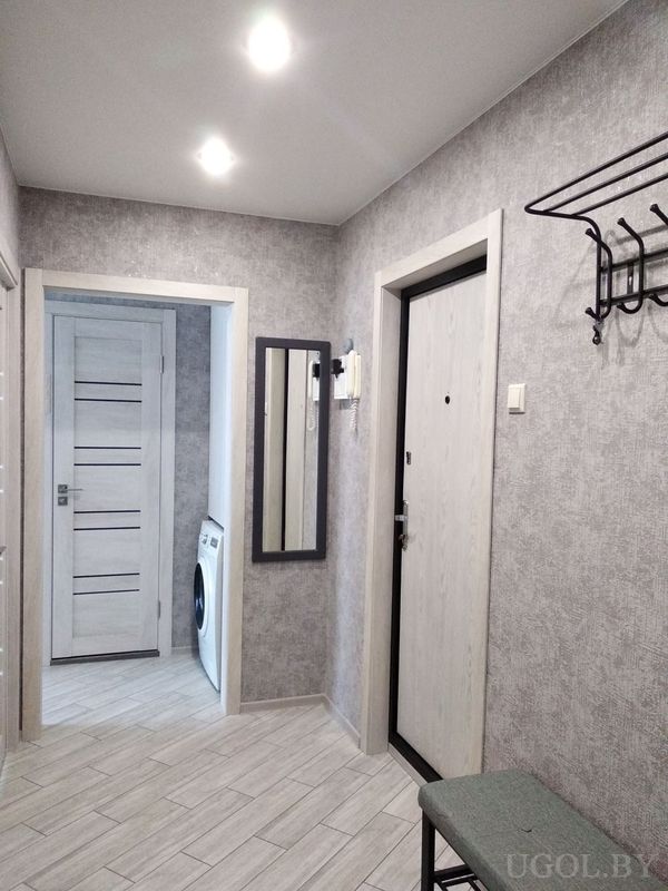 Современный коридор с серыми обоями, белыми дверями, зеркалом, вешалкой и скамьёй с зелёной подушкой.