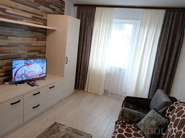 Современная гостиная с телевизором на светлой тумбе, стеной из разноцветных деревянных панелей, диваном и большим окном с белыми шторами.