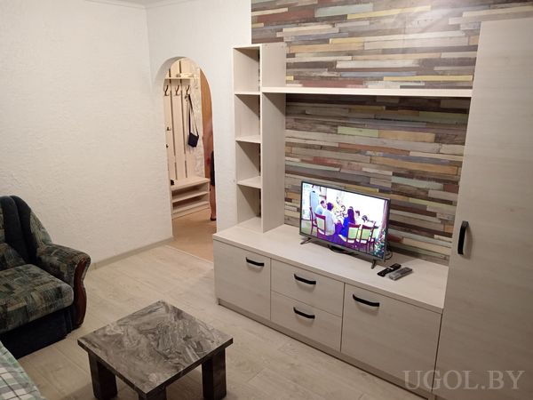 Уютная гостиная с белой тумбой и телевизором, разноцветной деревянной стеной, зелёным креслом и арочным проёмом в прихожую.