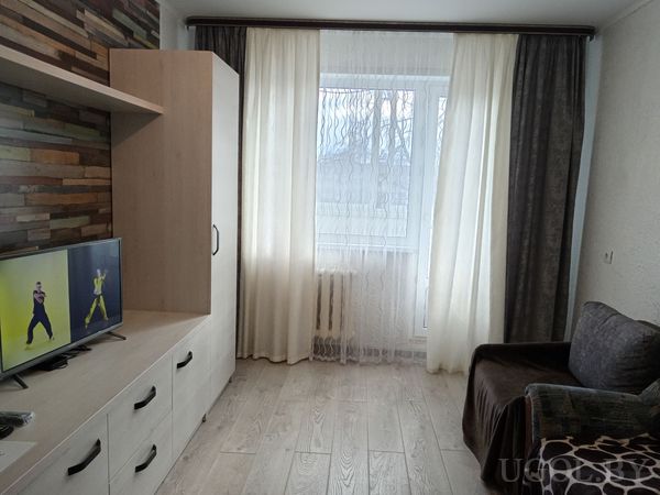 Светлая гостиная с телевизором на тумбе, стеной с декоративным камнем, тёмным диваном и большим окном с белыми шторами.