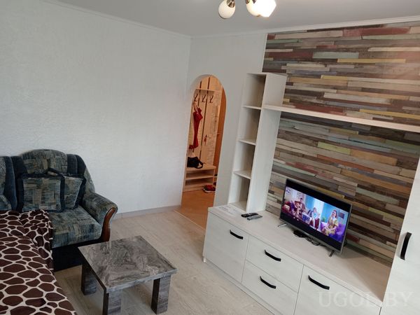 Комната с телевизором на белой тумбе, разноцветной деревянной стеной, креслом с узорчатой обивкой и журнальным столиком.