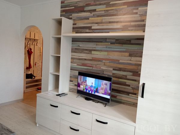 Современная гостиная с телевизором на светлой тумбе, разноцветной деревянной стеной и арочным проёмом в коридор.