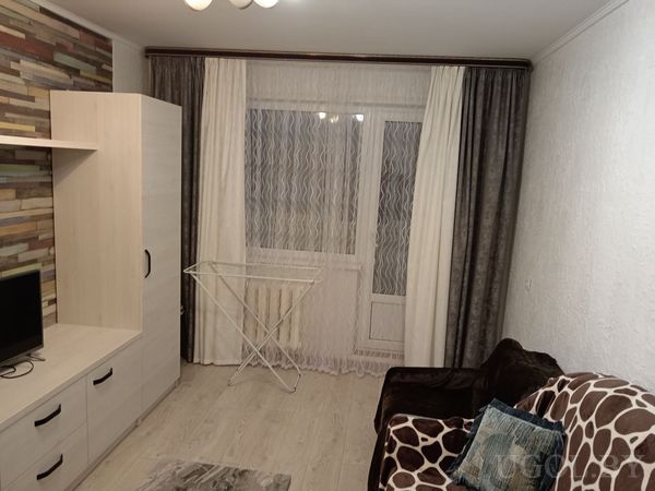 Небольшая гостиная с телевизором на светлой тумбе, разноцветной деревянной стеной, диваном и сушилкой для белья у окна.