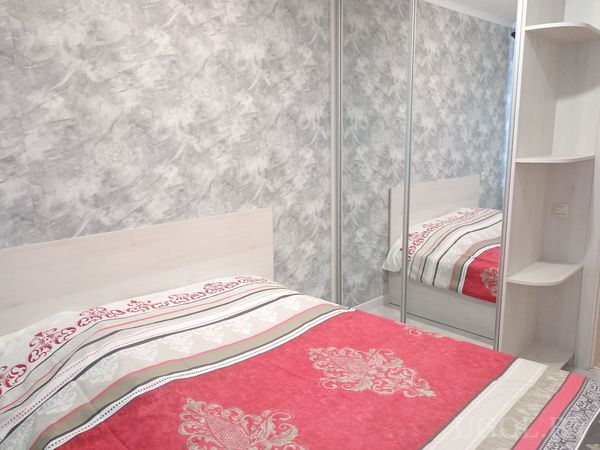 Современная спальня с белой кроватью и красно‑серым покрывалом, серой акцентной стеной и большим зеркальным шкафом.