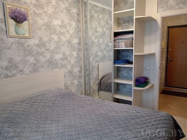 Светлая спальня с кроватью в сером покрывале, серой стеной с картиной лаванды и встроенными полками рядом с зеркальным шкафом.