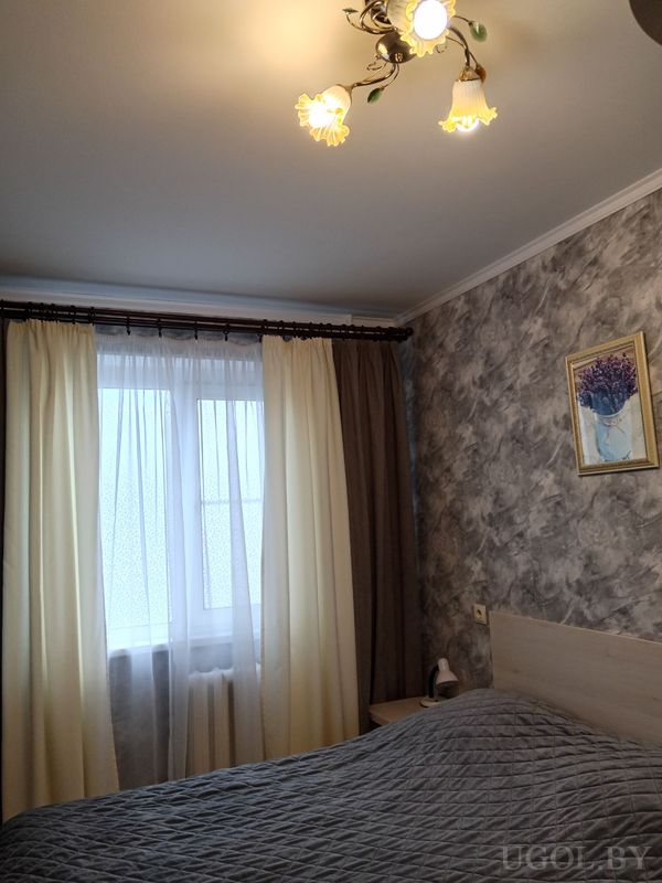 Уютная спальня с кроватью в сером покрывале, серой стеной с картиной фиолетовых цветов и окном с белыми и коричневыми шторами.