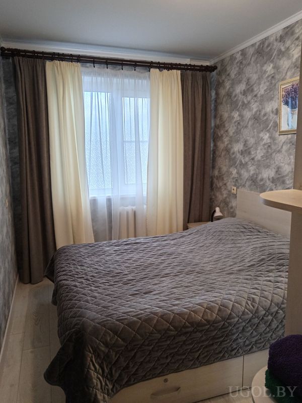 Современная спальня с кроватью в сером покрывале, светлым деревянным каркасом, серой стеной и окном с белыми и тёмными шторами.