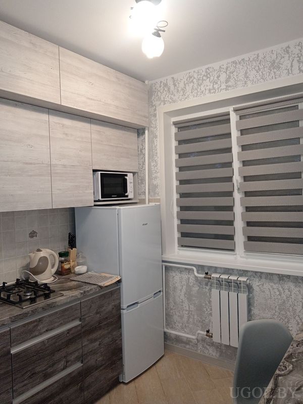 Компактная кухня с плитой, чайником, холодильником с микроволновкой сверху и окном с рулонной шторой.