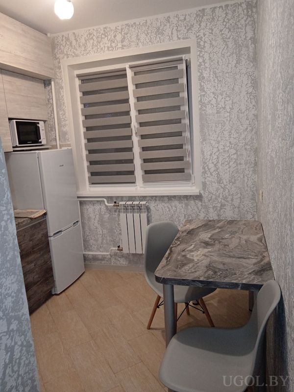 Небольшая кухня с холодильником и микроволновкой, окном с рулонной шторой и маленьким столиком с двумя стульями.