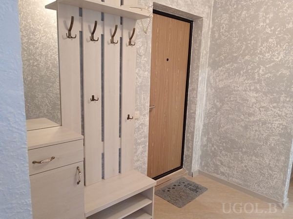 Светлая прихожая с деревянной вешалкой с крючками, тумбой для обуви и входной дверью с ковриком.