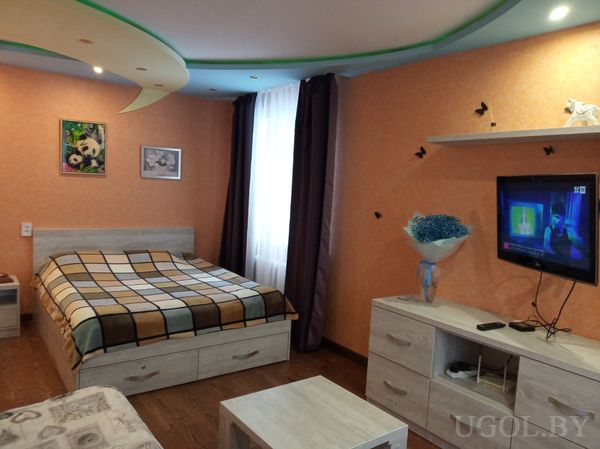 Современная спальня с двуспальной кроватью в клетчатом покрывале, телевизором на стене, комодом с вазой синих цветов и оранжевой стеной.