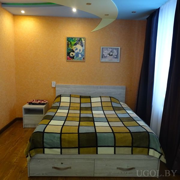 Уютная спальня с кроватью в клетчатом покрывале, светлым каркасом с ящиками, прикроватной тумбой и оранжевой стеной с картинами.